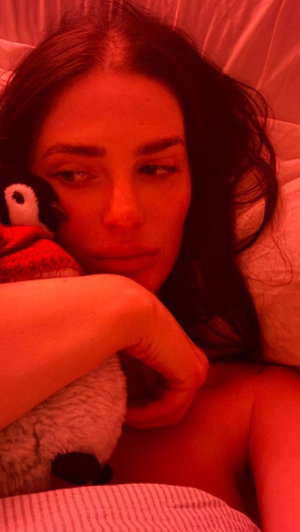 Woman with dark hair lying in bed, holding a stuffed penguin, red lighting.