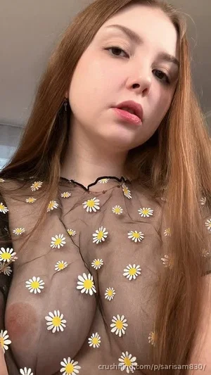 Woman with long brown hair in sheer floral top, indoors, looking at camera.
