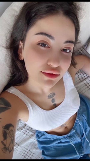Woman with tattoos wearing white tank top and blue shorts, lying on bed.