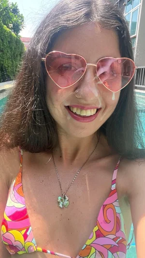 Woman with dark hair and heart-shaped sunglasses in colorful bikini by pool.