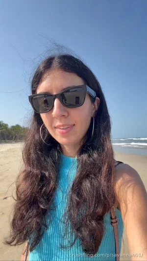 Woman with long dark hair wearing sunglasses and a blue top at the beach.