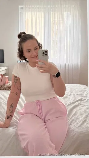 Woman with tattoos in white top and pink pants sitting on bed, taking mirror selfie.
