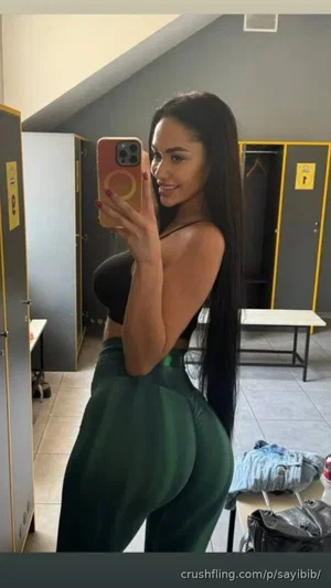 Woman with long dark hair in black crop top and green pants, taking mirror selfie in locker room.