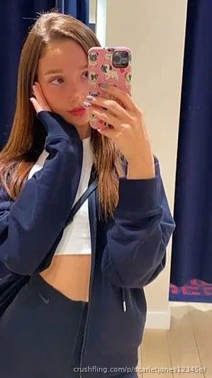 Woman in navy jacket and white crop top taking mirror selfie indoors.