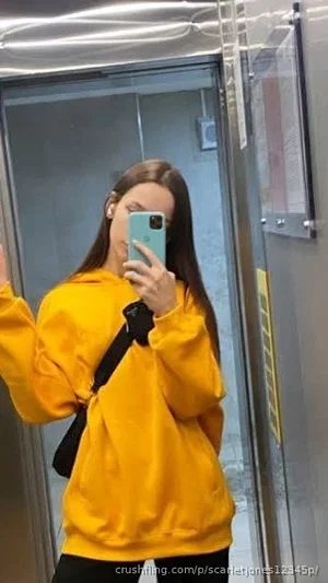 Woman in yellow hoodie and black pants takes mirror selfie in elevator.