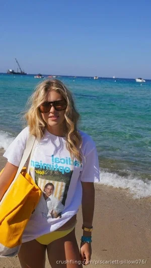 Blonde woman in white t-shirt and yellow bikini bottoms at beach, carrying yellow bag.