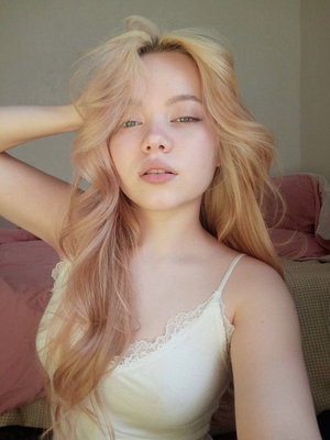 Woman with blonde hair in a white lace camisole, posing indoors near a bed.