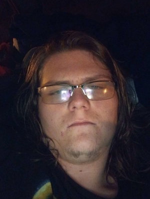 Person with long dark hair and glasses wearing a dark shirt in a dimly lit indoor setting.