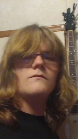 Person with long blonde hair and glasses wearing black top indoors.