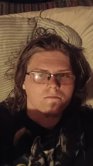 Person with long hair and glasses lying on a bed, wearing a dark shirt.