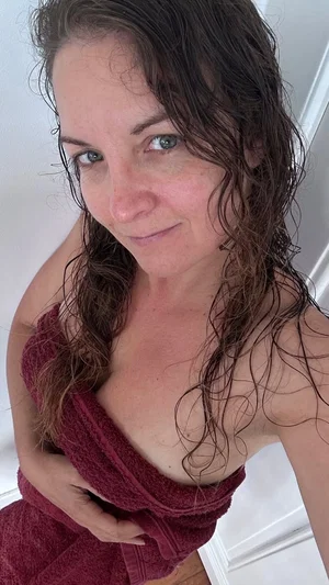 Woman with wet hair wrapped in a maroon towel, indoors, smiling at camera.
