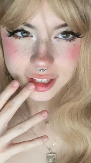 Blonde woman with makeup and nose ring, hand near lips, wearing necklace.