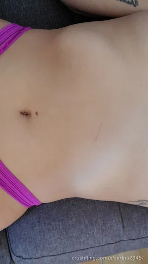 Woman in purple bikini top lying on gray couch, showing stomach and tattoo.