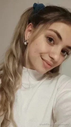 Woman with blonde wavy hair in a high ponytail, wearing a white turtleneck sweater, smiling at the