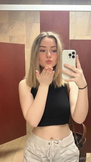 Woman with blonde hair in black crop top and beige pants, posing in mirror selfie indoors.