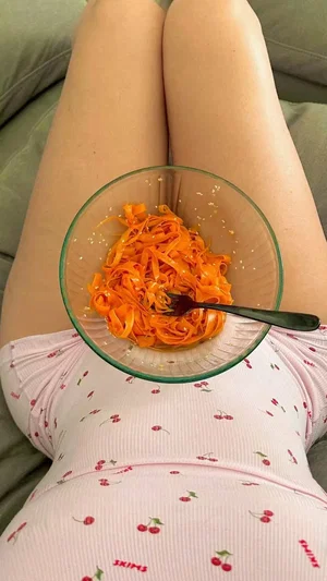 Woman in pink cherry-patterned top holding bowl of carrot salad on lap, sitting on couch.