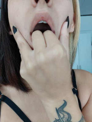 Close-up of woman with fingers in mouth, black nails, wrist tattoo.