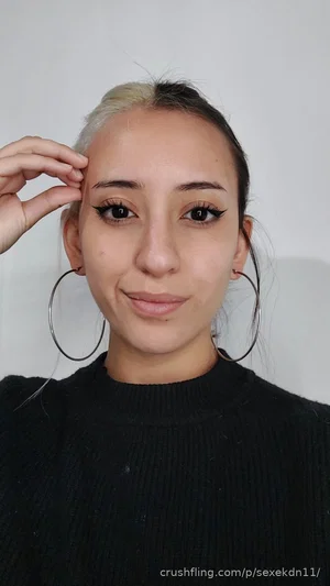 Woman with two-toned hair, wearing black sweater and large hoop earrings, posing indoors.
