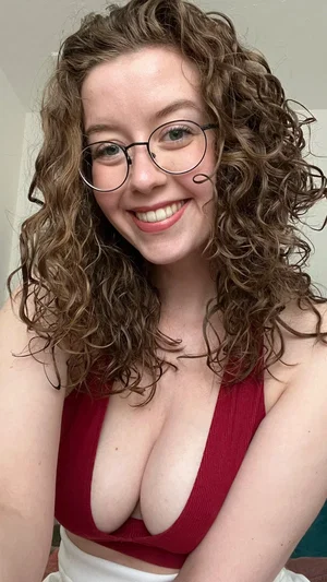Woman with curly hair and glasses wearing a red top indoors.