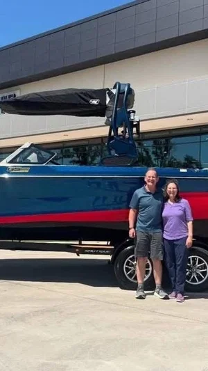 A man and woman stand in front of a blue and red speedboat on a trailer outside a building.