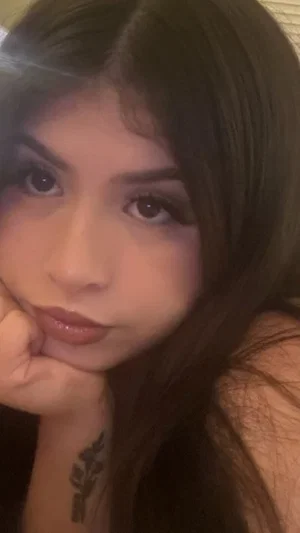 Woman with long dark hair and makeup resting chin on hand, tattoo on wrist, indoor setting.