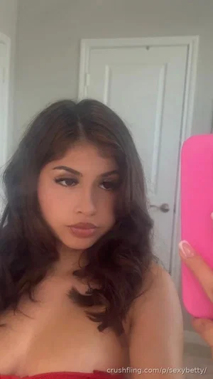 Woman with dark wavy hair in red strapless top, holding pink phone, mirror selfie in indoor setting.
