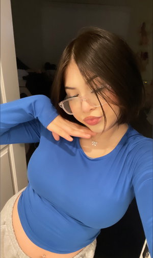 Woman with short dark hair wearing blue crop top and glasses, posing indoors.