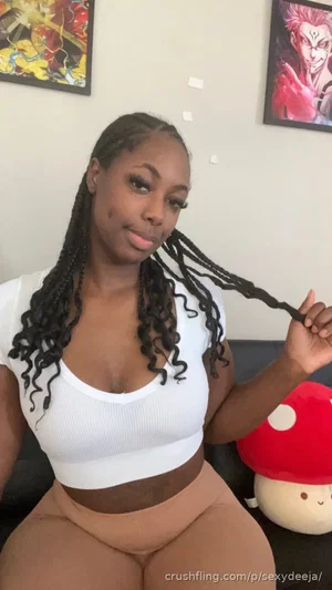 Woman with braided hair in white crop top and beige pants, sitting indoors near anime posters.