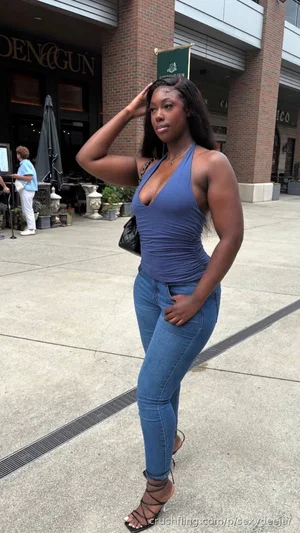 Woman in blue tank top and jeans posing outdoors on a sidewalk.