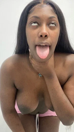 Woman with long black hair in pink underwear posing indoors, hand on neck, tongue out.