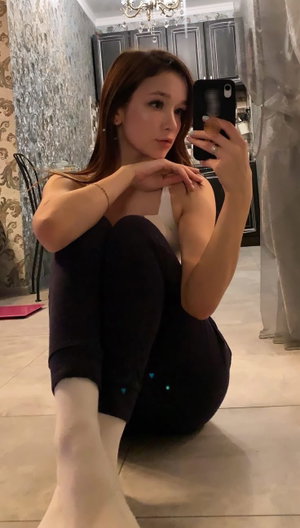 Brunette woman in white top and black pants taking indoor selfie.