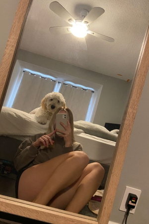 Woman in long-sleeve top and underwear takes mirror selfie in bedroom with dog on bed.