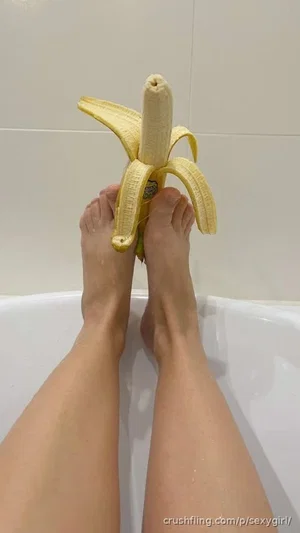Person's bare legs and feet in a bathtub holding a peeled banana.