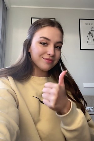 Woman with long brown hair in a beige sweatshirt giving a thumbs-up indoors.