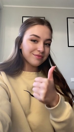 Woman with long brown hair in a beige sweatshirt giving a thumbs-up indoors.