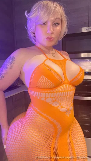 Woman with short blonde hair in orange mesh bodysuit, standing in kitchen with purple lighting.