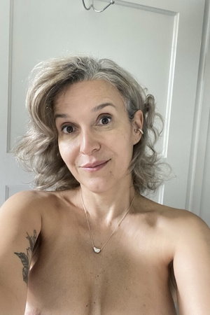 Woman with gray curly hair, bare shoulders, necklace, tattoo on arm, indoor setting.