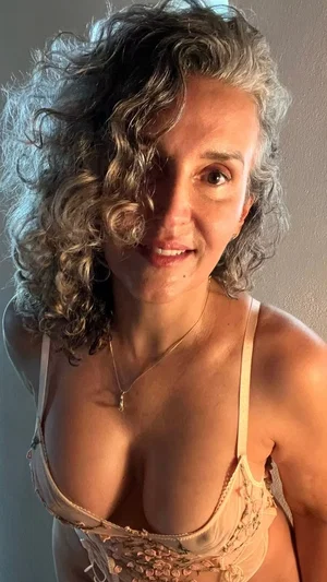 Woman with curly gray hair in beige lace lingerie, smiling at camera indoors.