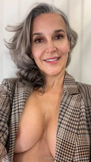 Woman with gray hair, bare chest, wearing open plaid blazer, indoor setting.