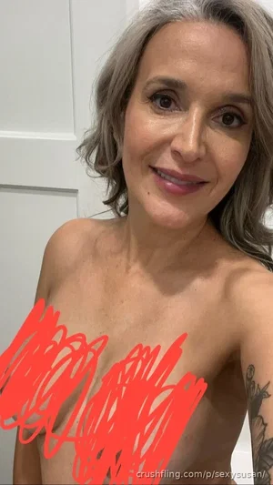 Woman with gray hair and tattoo, topless with red scribble over chest, indoors.