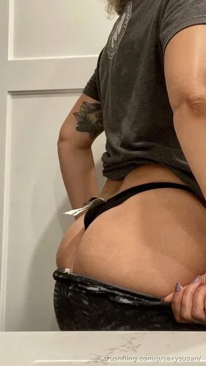 Woman in black thong and gray t-shirt, showing backside, indoor setting.