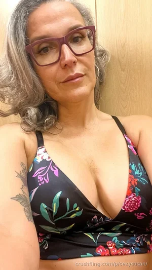 Woman with gray hair and glasses wearing floral bra, indoor selfie.