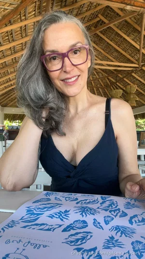 Woman with gray hair and purple glasses wearing a dark blue top, sitting at a table under a