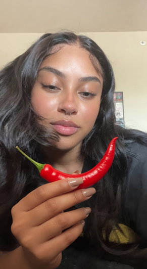 Woman with long dark hair holding a red chili pepper indoors.