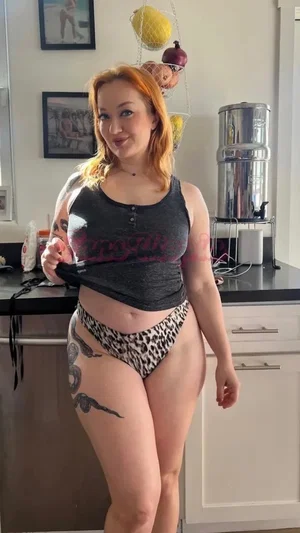 Woman with red hair and tattoos wears a dark top and leopard-print underwear in a kitchen.
