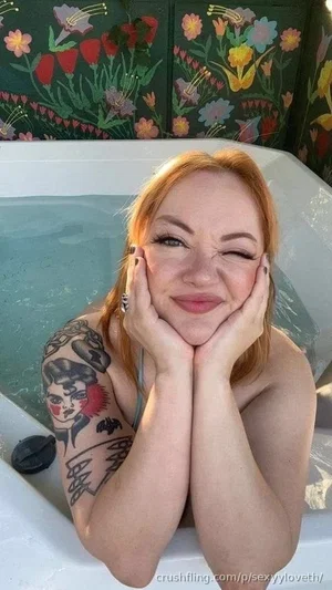 Woman with red hair and tattooed arm in a hot tub, smiling with hands on face.