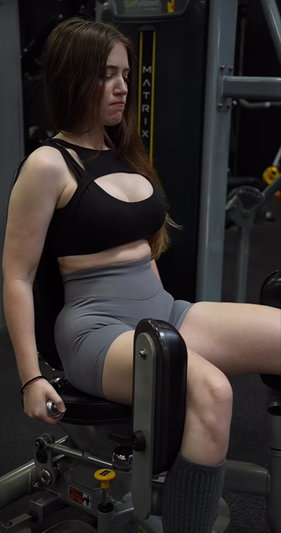 Woman in black sports bra and gray shorts using gym machine, indoor setting.