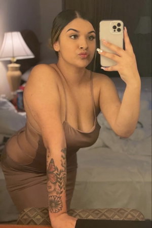 Woman in sheer brown dress taking mirror selfie in bedroom.