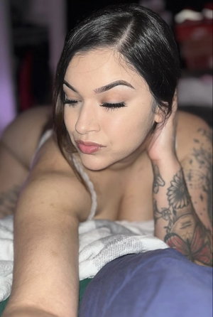 Woman with dark hair and tattoos lying in bed wearing white top.