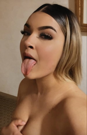 Woman with blonde hair and makeup sticking tongue out indoors.
